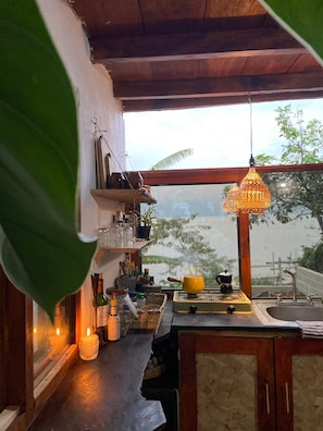 Cozinha com vista pela janela, luminária pendente e diversos utensílios.