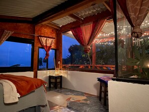 Quarto com cama, uma mesinha, janela com cortinas e vista para o exterior.