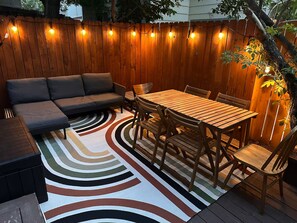 Eine Holzterrasse mit einer Couch, einem Esstisch und Stühlen, umgeben von einem Holzzaun und Lichterketten.