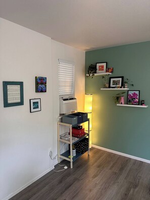 Ein Zimmer mit weißer Wand, einer Türkis-farbenen Akzentwand, Holzboden, einem weißen Regal mit verschiedenen Gegenständen, einem Fenster mit Jalousien und einer Lampe.