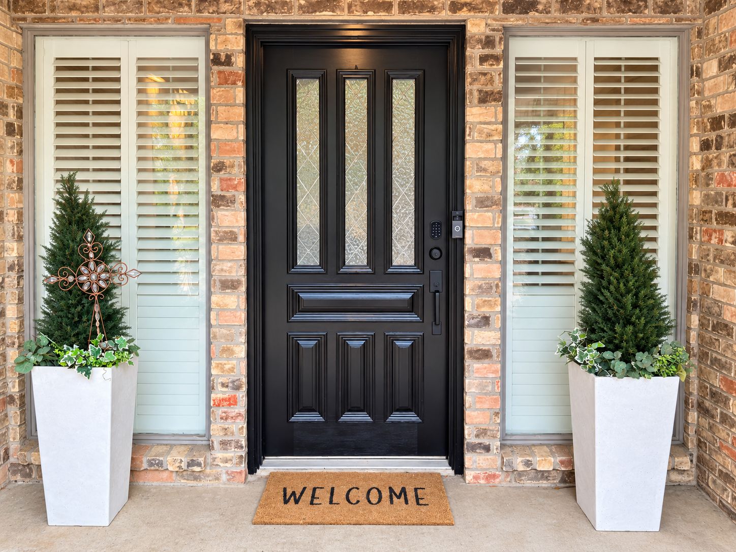 A black front door with a welcome mat, flanked by two white planters with greenery.