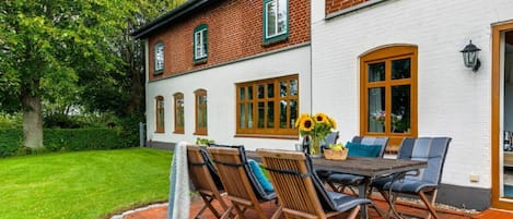 Une maison en briques avec une façade blanche, une terrasse avec une table et des chaises, et un jardin avec un arbre.