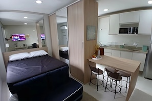 A bedroom with a bed, a desk with two chairs, a mirror, and a kitchenette. - SHB - Apê Charmoso no Embaré (Santos)