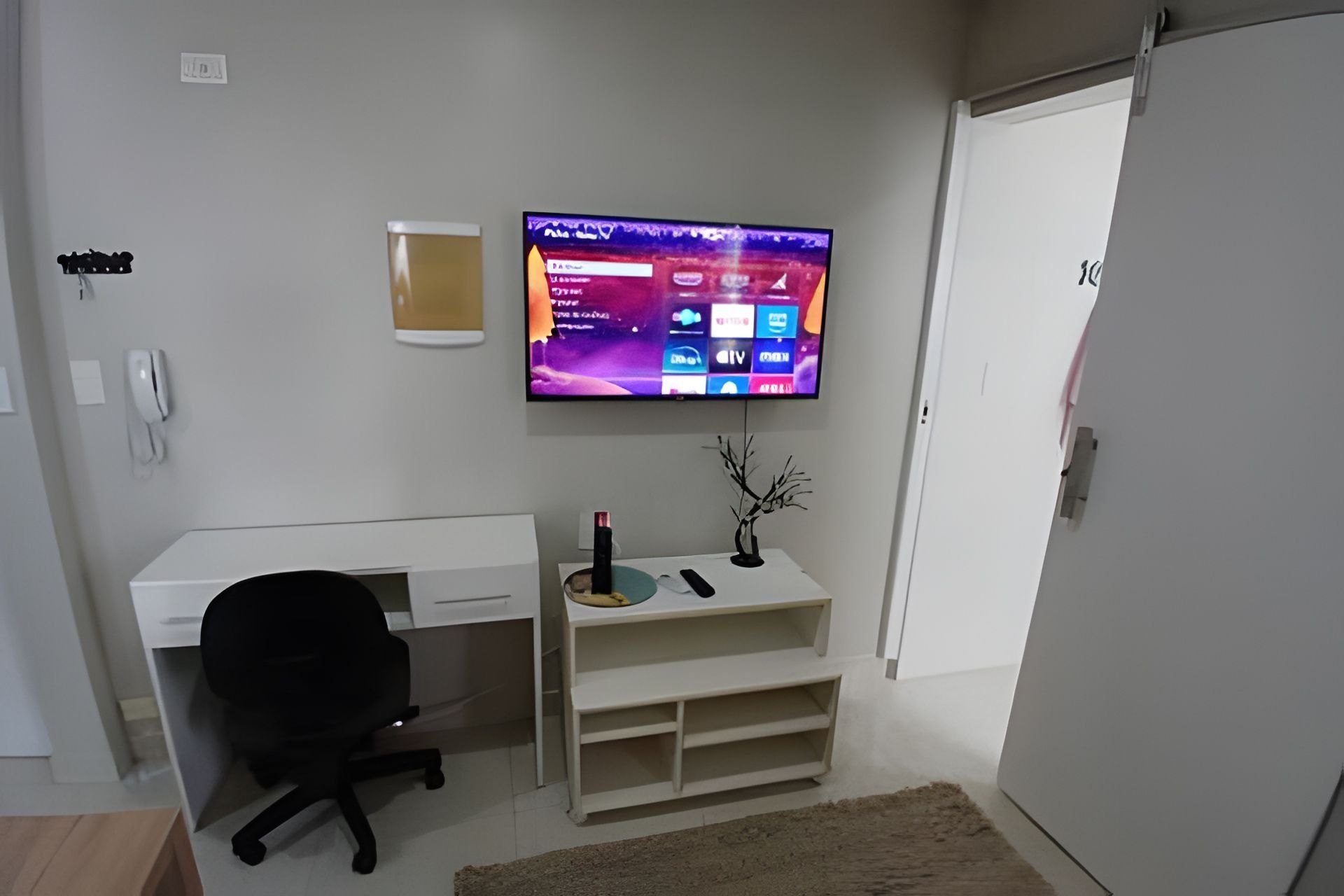 Quarto moderno com televisão de tela plana na parede, escrivaninha branca com cadeira, prateleira pequena com objetos decorativos e porta branca.