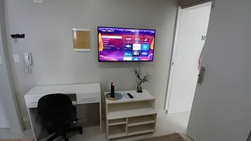 Quarto moderno com televisão de tela plana na parede, escrivaninha branca com cadeira, prateleira pequena com objetos decorativos e porta branca.