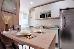 A modern kitchen with white cabinets, a stainless steel refrigerator, and a dining area with a wooden table set for two. - SHB - Apê Charmoso no Embaré (Santos)