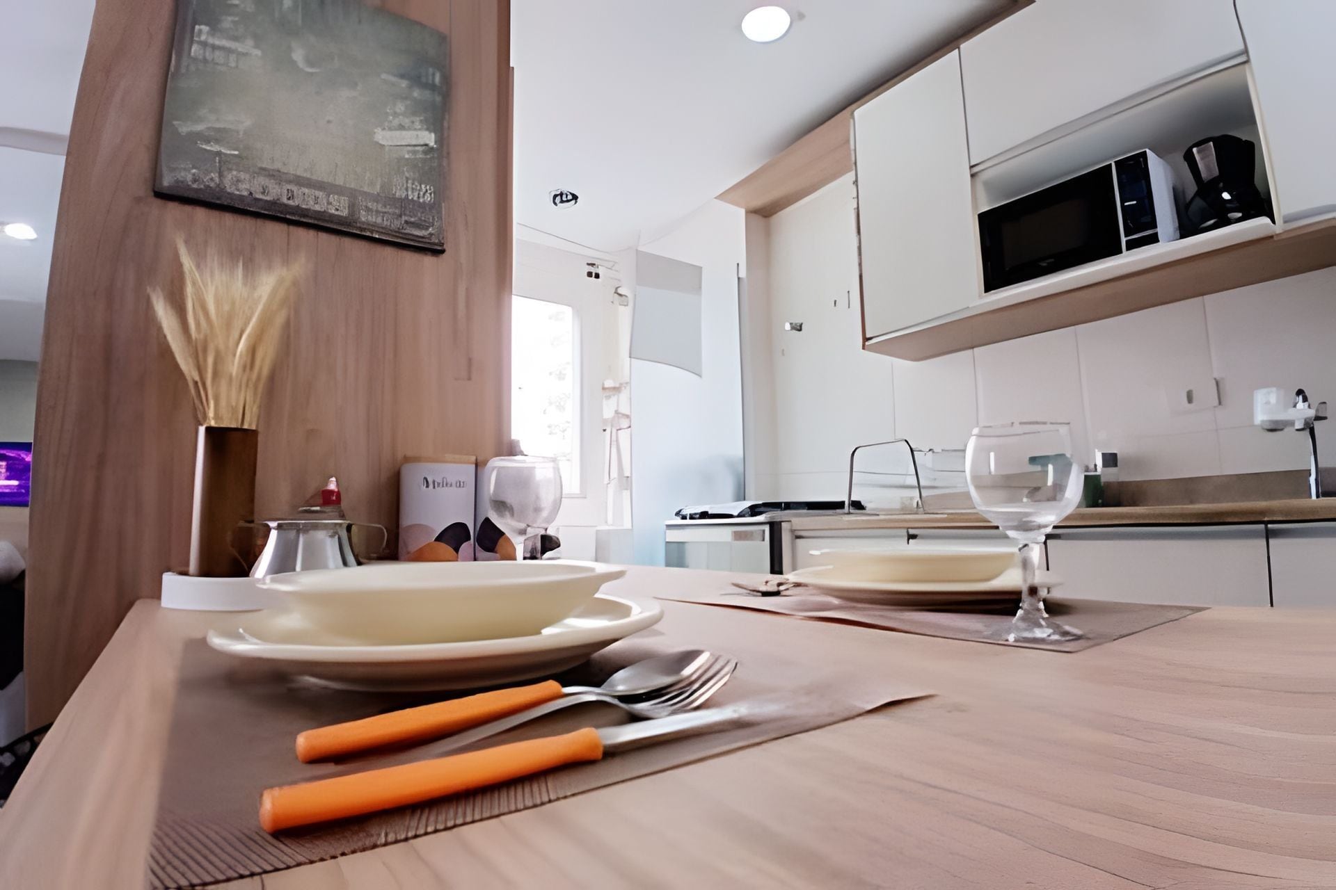 Una cocina moderna con armarios blancos, un microondas y una mesa de comedor dispuesta con platos, cubiertos y un jarrón de flores secas.