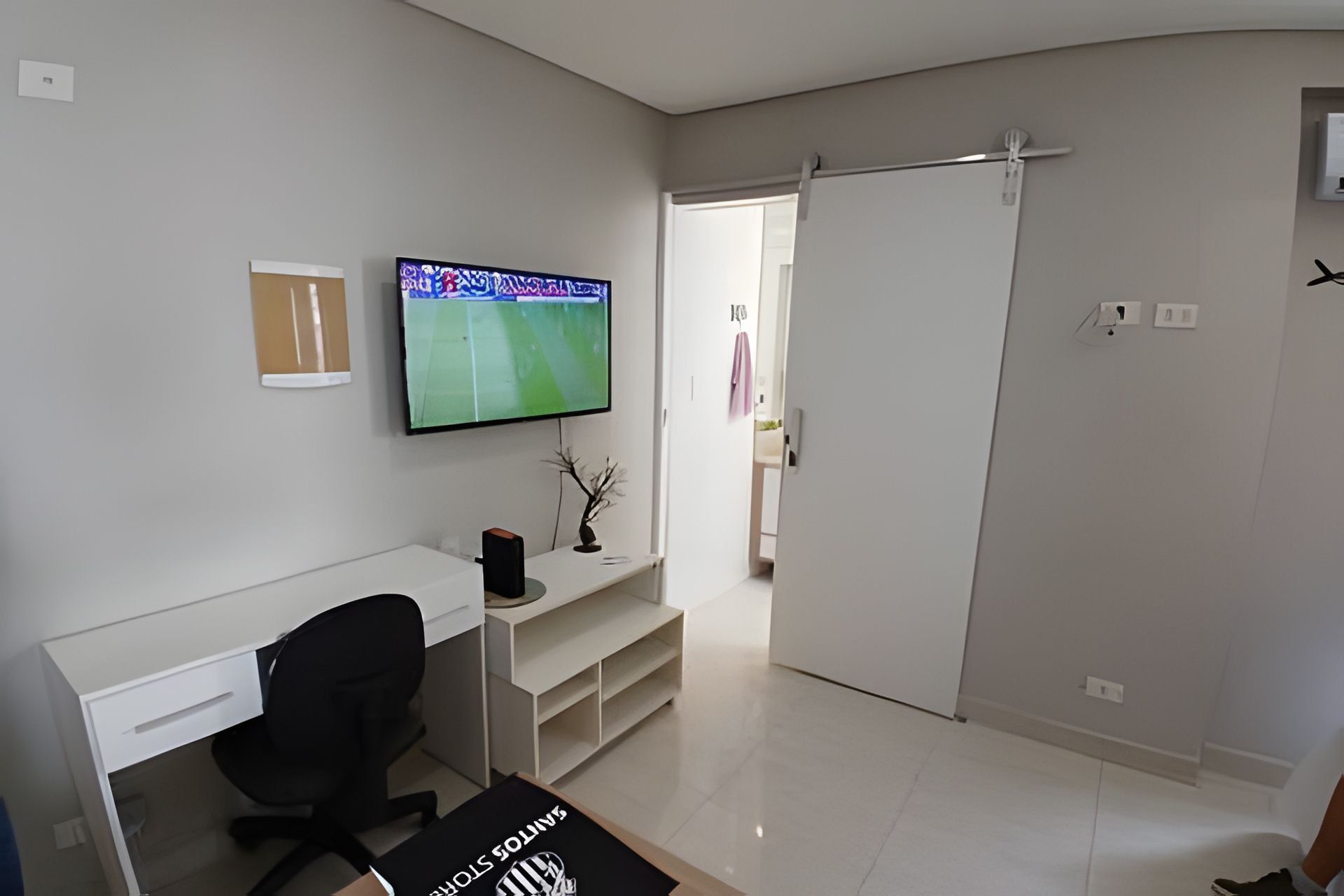 Una habitación moderna y minimalista con un escritorio blanco, una silla de oficina negra, un televisor de pantalla plana montado en la pared y una puerta corredera que da a otra habitación.
