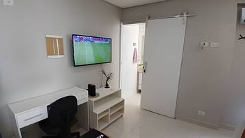 Una habitación moderna y minimalista con un escritorio blanco, una silla de oficina negra, un televisor de pantalla plana montado en la pared y una puerta corredera que da a otra habitación.