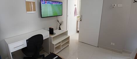 A modern, minimalistic room with a white desk, a black office chair, a flat-screen TV mounted on the wall, and a sliding door leading to another room.