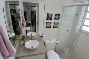A bathroom with a white sink, toilet, and a glass shower enclosure. There are four wall-mounted pictures. - SHB - Apê Charmoso no Embaré (Santos)