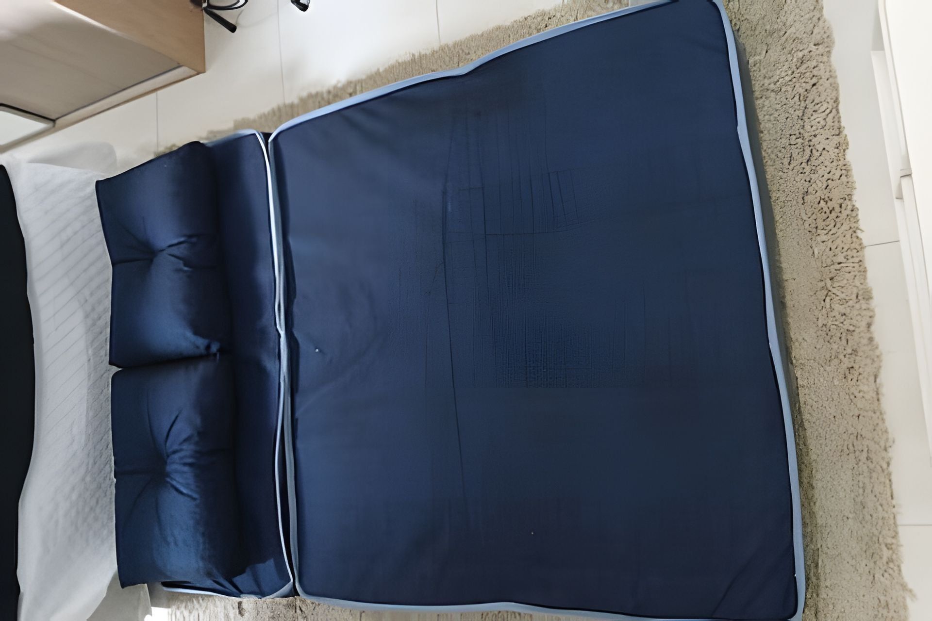 Una cama con un colchón y almohadas azul oscuro, un cabecero blanco y una alfombra texturizada en el suelo.