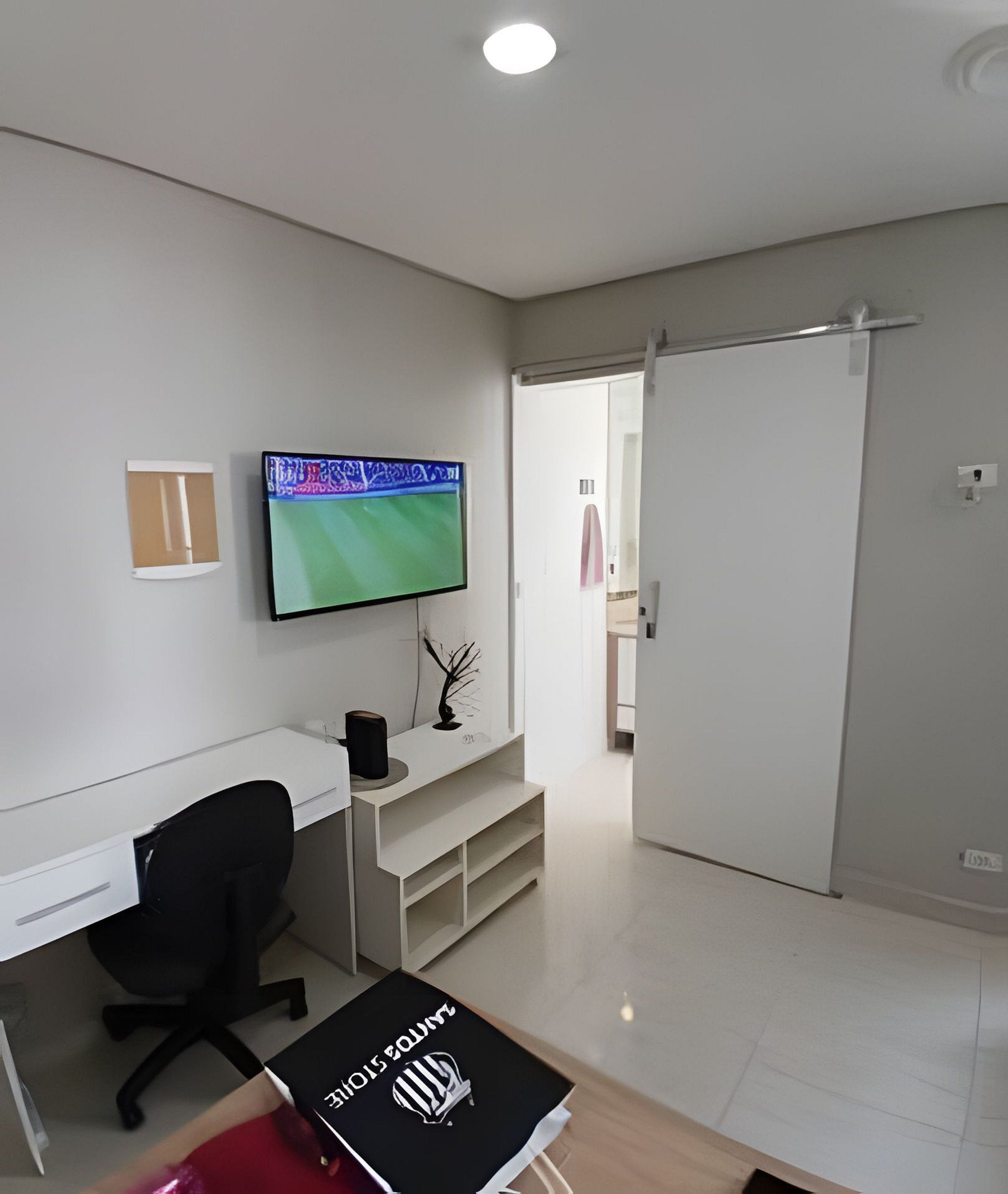 A modern, minimalistic office space with a white desk, black office chair, and a flat-screen TV mounted on the wall.