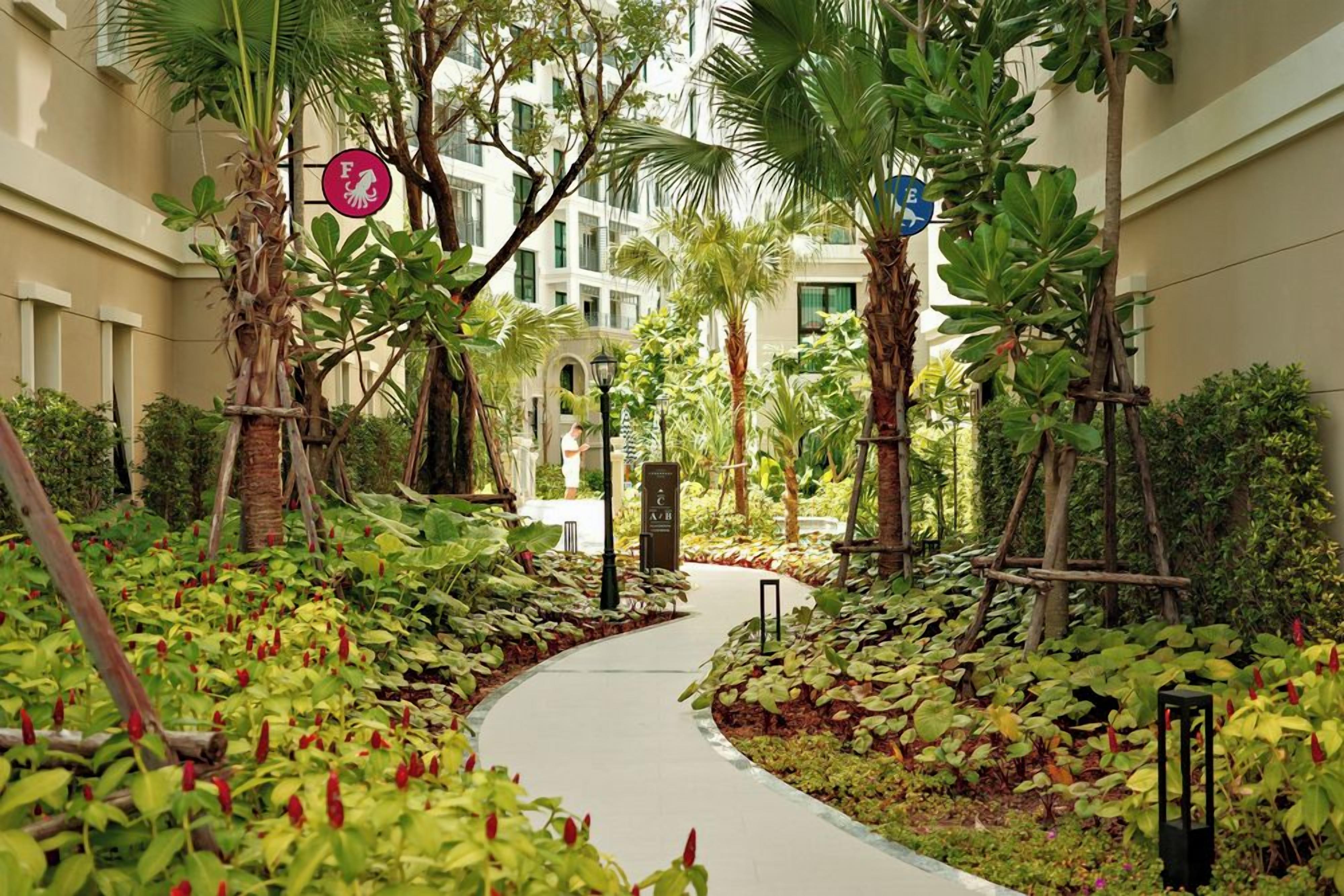 Ein Weg, der von üppiger Vegetation und Palmen gesäumt wird, führt zu einem Gebäude mit weißer Fassade und zahlreichen Fenstern.