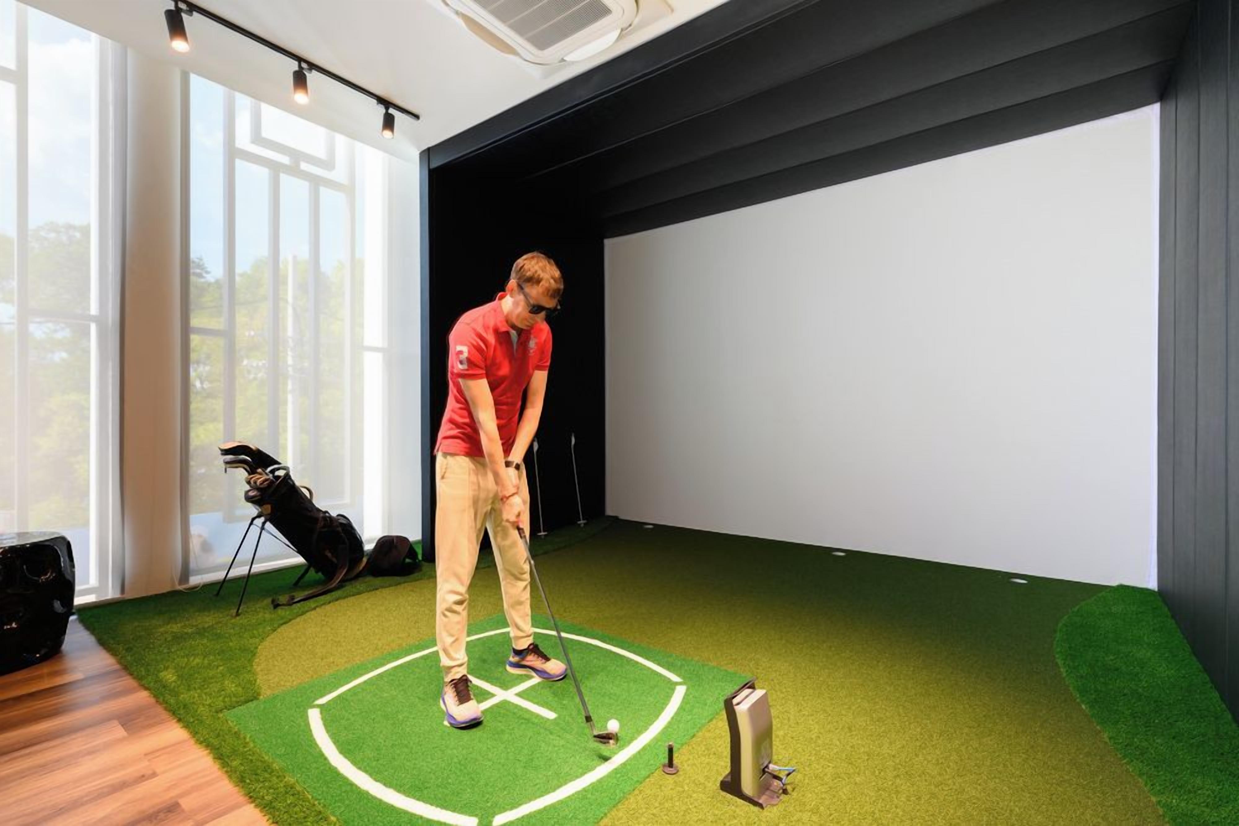 En person i en röd tröja som spelar golf inomhus, med en grön konstgräsmatta som underlag och en vit skärm i bakgrunden.