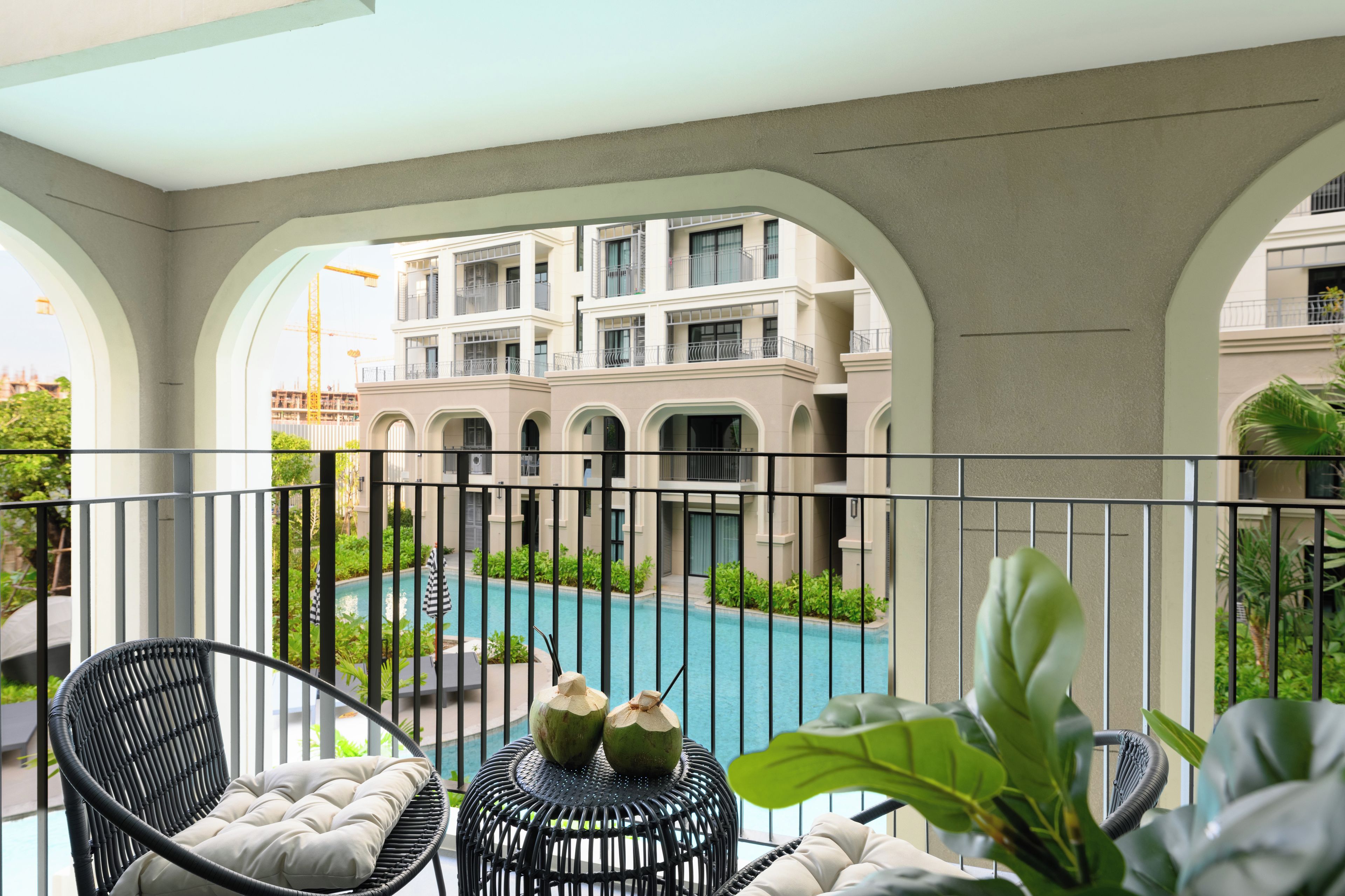 A balcony with a view of a swimming pool, a lounge chair, and a table with coconuts.