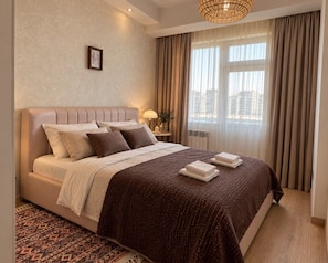 Ein Hotelzimmer mit einem großen Bett, einem Nachttisch mit Lampe, Blick auf die Stadt durch das Fenster und einem gemusterten Teppich.