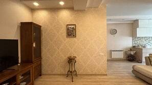 Een woonkamer met een televisie, een houten kast, behang met een patroon, een kleine tafel met een kaars, en een keukenhoek met een klok en een radiator.