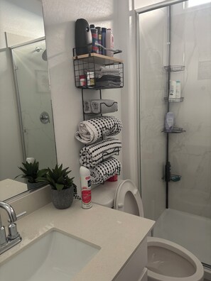 Un baño con cabina de ducha de vidrio, inodoro, lavabo con espejo y toalleros con toallas dobladas y artículos de aseo.