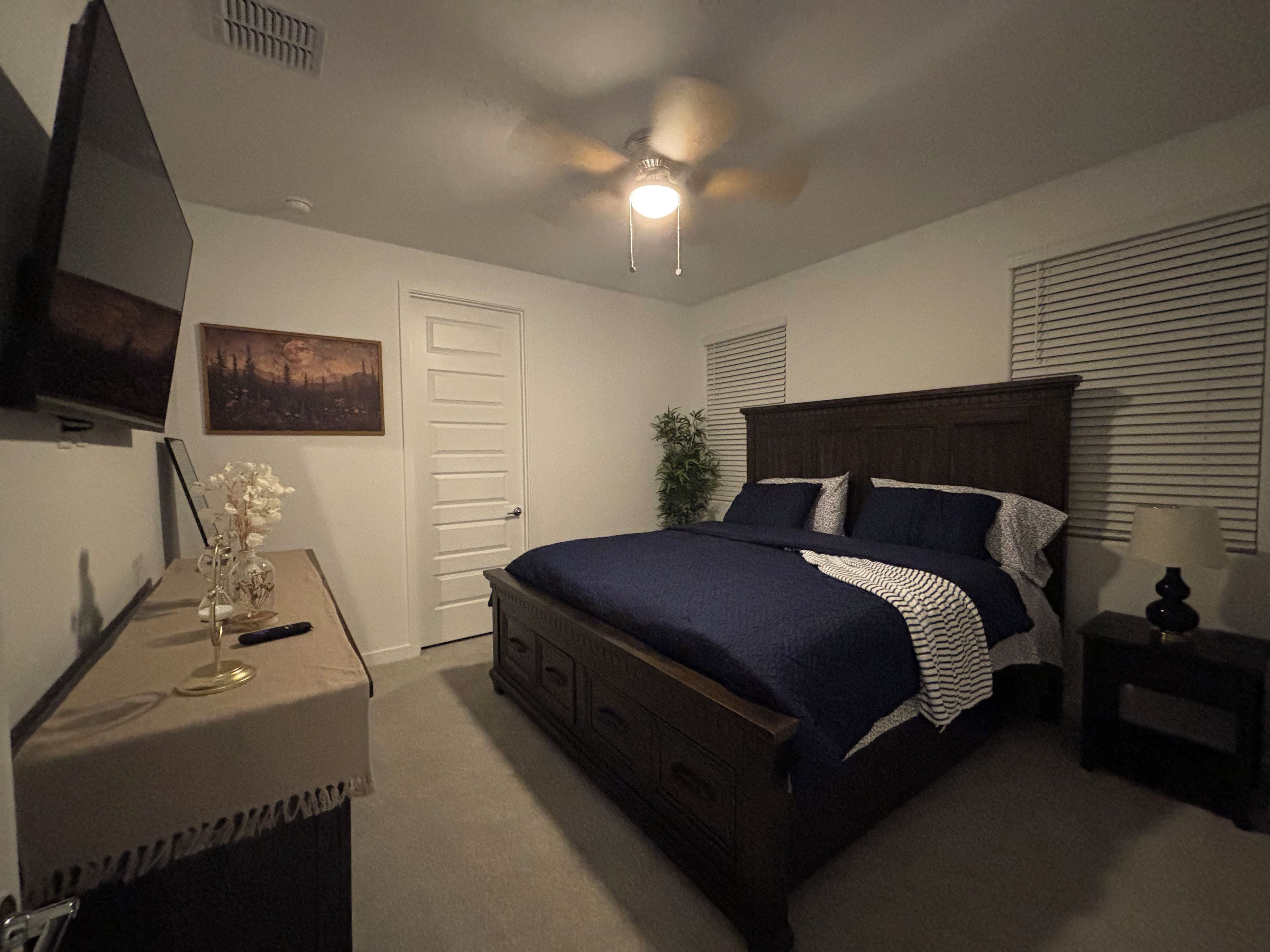 A bedroom with a bed, a desk, a chair, a ceiling fan, a window with blinds, and a painting on the wall.