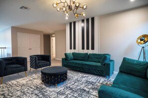 A modern living room with a green sofa, black armchairs, a round ottoman, and a unique chandelier. - Luxury 9BR Home Near Disney | Solara Resort 81SA (Kissimmee)