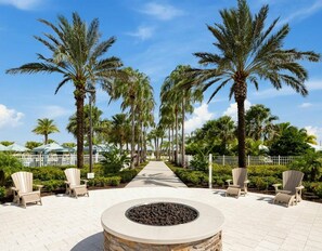A pathway lined with palm trees leading to a seating area with Adirondack chairs and a central fire pit. - Luxury 9BR Home Near Disney | Solara Resort 81SA (Kissimmee)