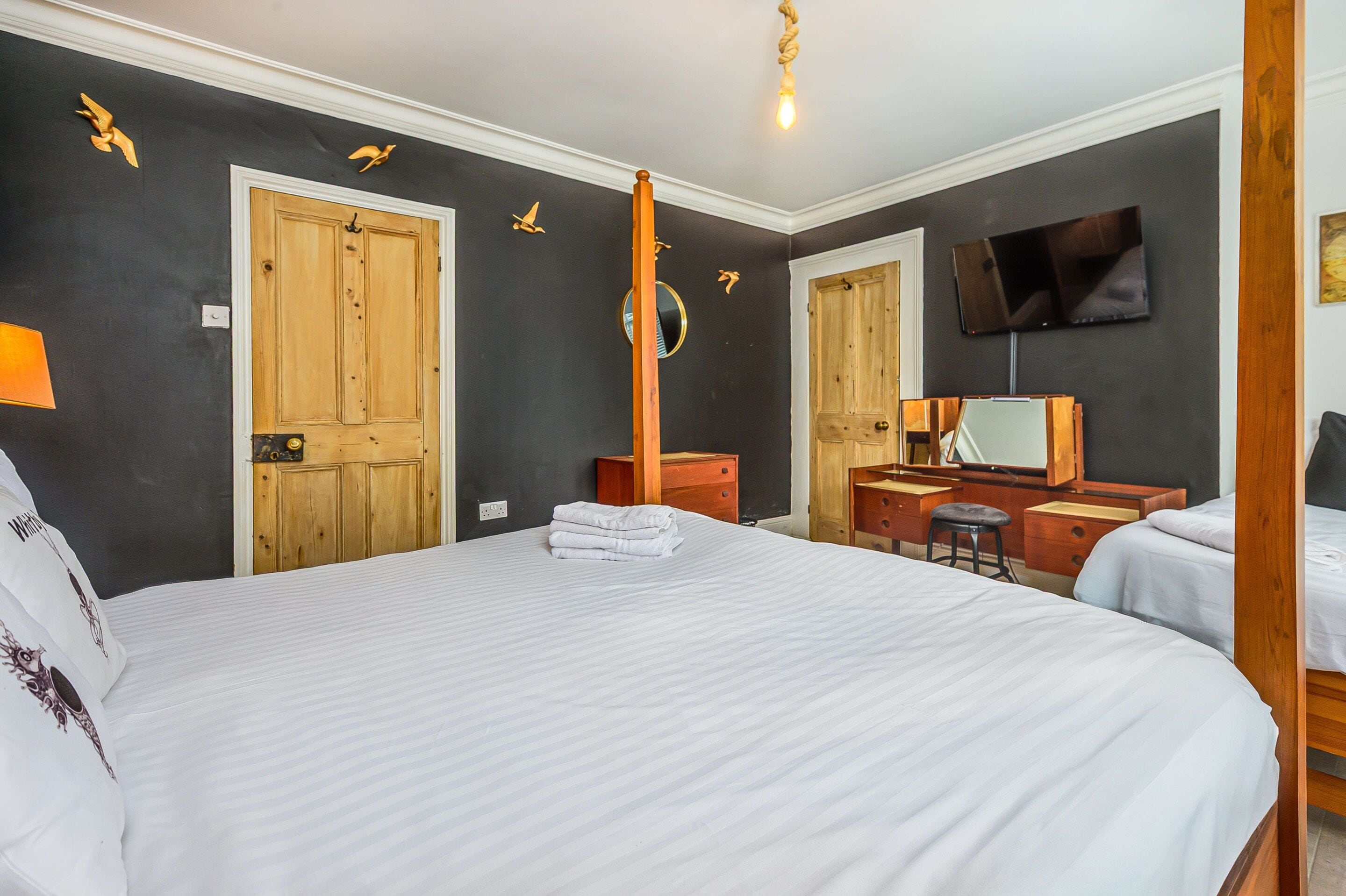 A hotel room with a large bed, a wooden wardrobe, a desk with a chair, a television mounted on the wall, and a ceiling light fixture.