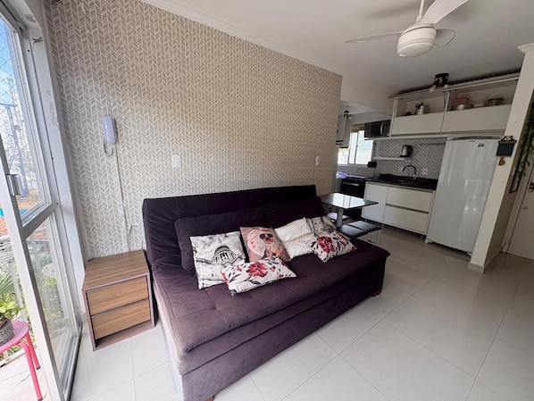 A modern living room with a sofa, decorative pillows, a small side table, and a kitchen area with cabinets and appliances. - Vrbo Property (Balneário Camboriú)