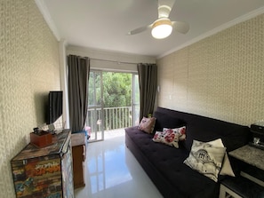 A living room with a black sofa, a flat-screen TV, a ceiling fan, and a sliding glass door leading to a balcony. - Vrbo Property (Balneário Camboriú)