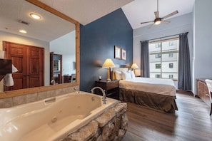 Quarto de hotel com uma banheira grande, cama, mesa de cabeceira com abajures e vista para um edifício pela janela.
