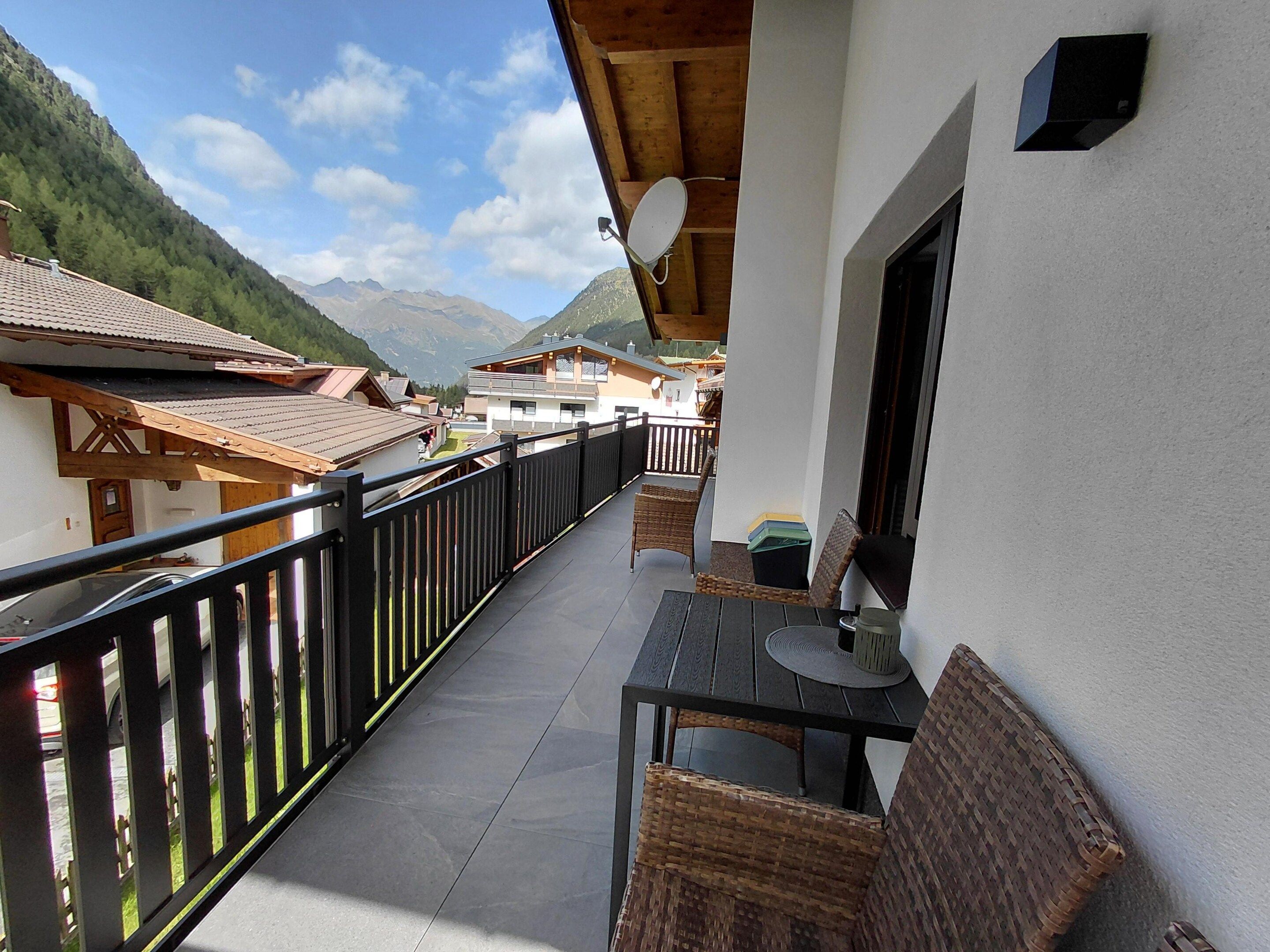A balcony with a view of a mountain landscape, a table with chairs, and a small table with a cup.
