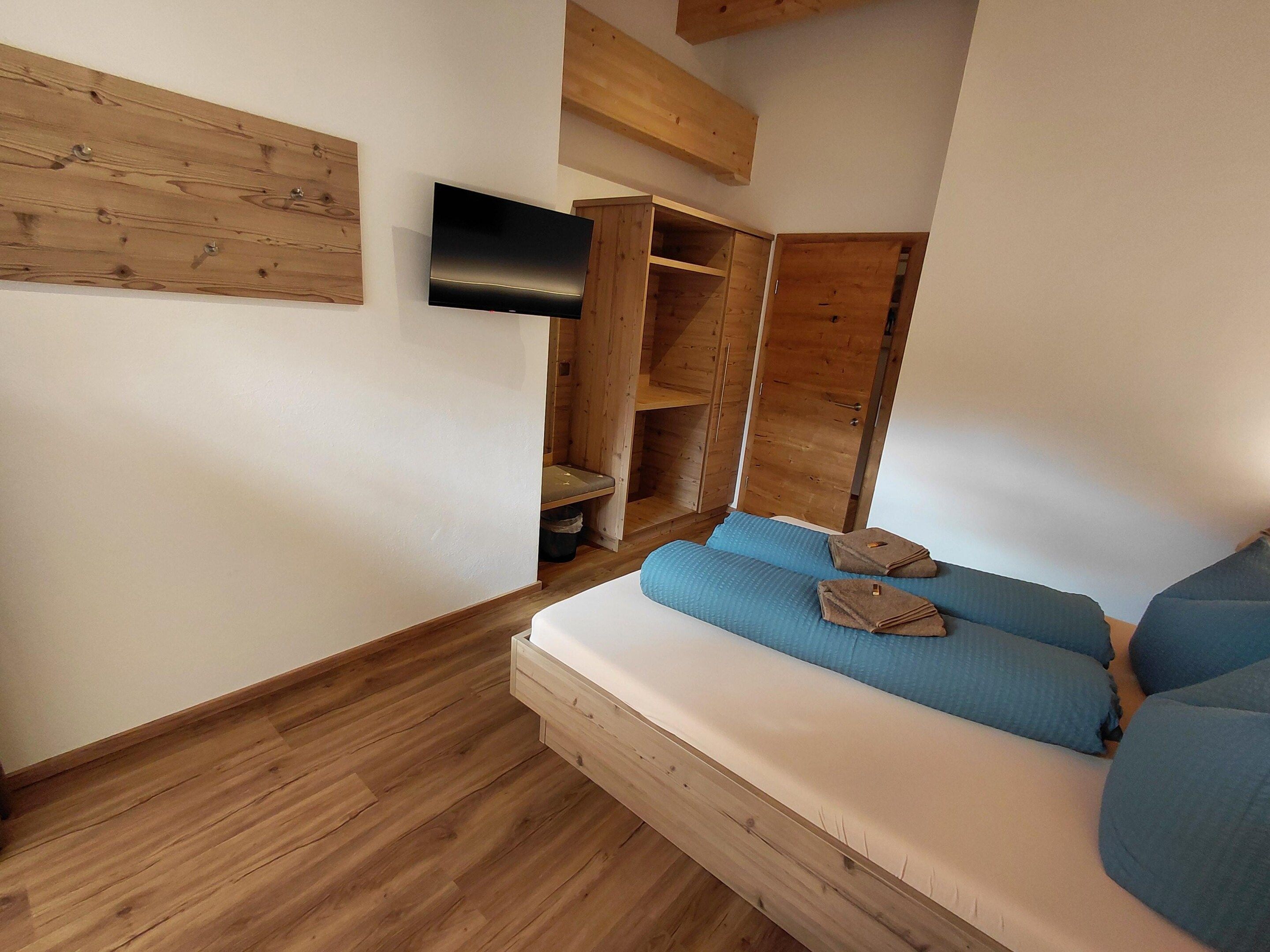A bedroom with a bed, a TV mounted on the wall, wooden shelving, and a wooden floor.