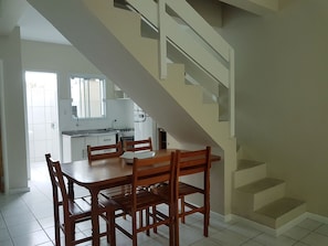 Un área de comedor con una mesa y sillas de madera, una cocina al fondo y una escalera que lleva a un nivel superior.