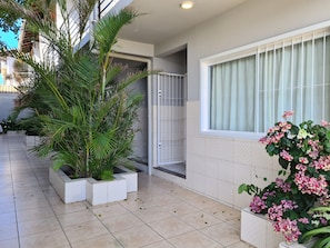 Un área exterior moderna con suelo de baldosas, plantas en macetas, un ventanal de vidrio que da a un espacio cubierto y una ventana con cortinas.