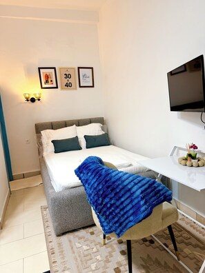 A bedroom with a bed, a desk, a chair, a TV, and wall-mounted lights. - Cozy studio apartment in Milan, in the heart of Bonapriso (Douala)