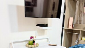 Una camera d'albergo con una scrivania bianca, una televisione fissata al muro, una sedia con una coperta blu e un armadio con vestiti piegati.