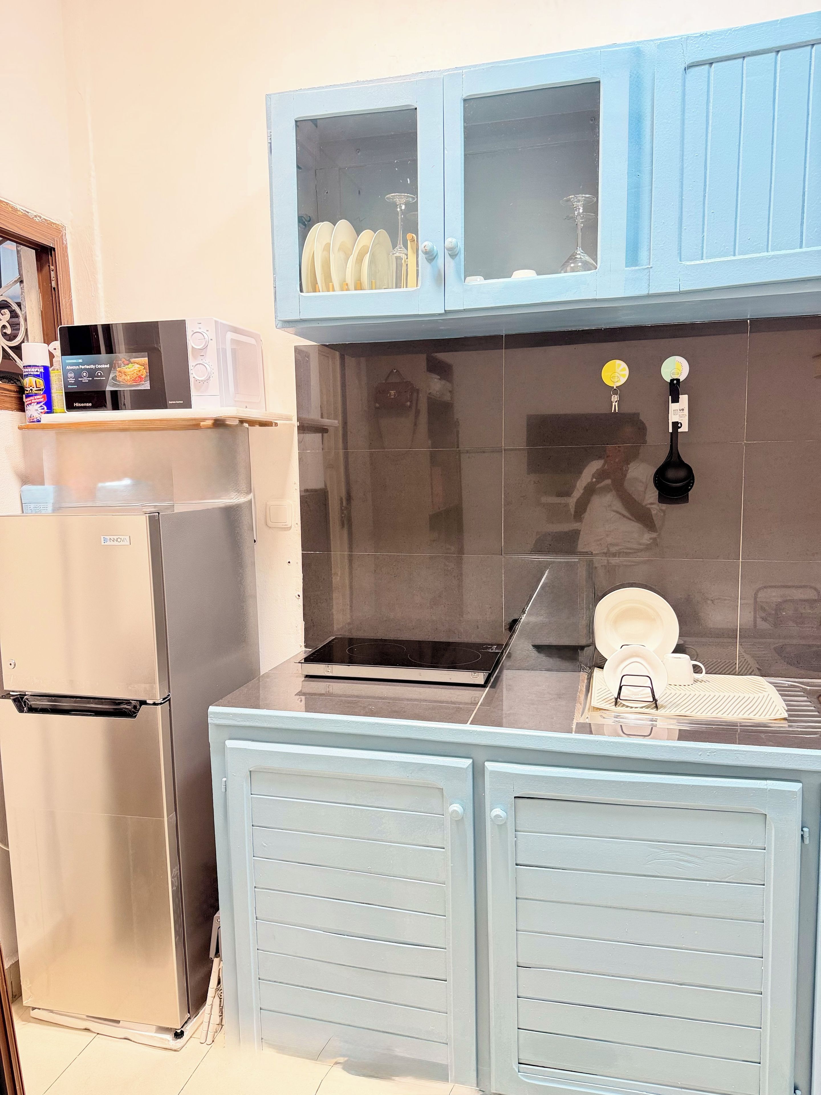 Una cucina compatta con frigorifero in acciaio inossidabile, forno a microonde e piano cottura a induzione nero. Sopra il piano cottura, armadietti blu con vetri, e un piccolo scolapiatti sul ripiano.