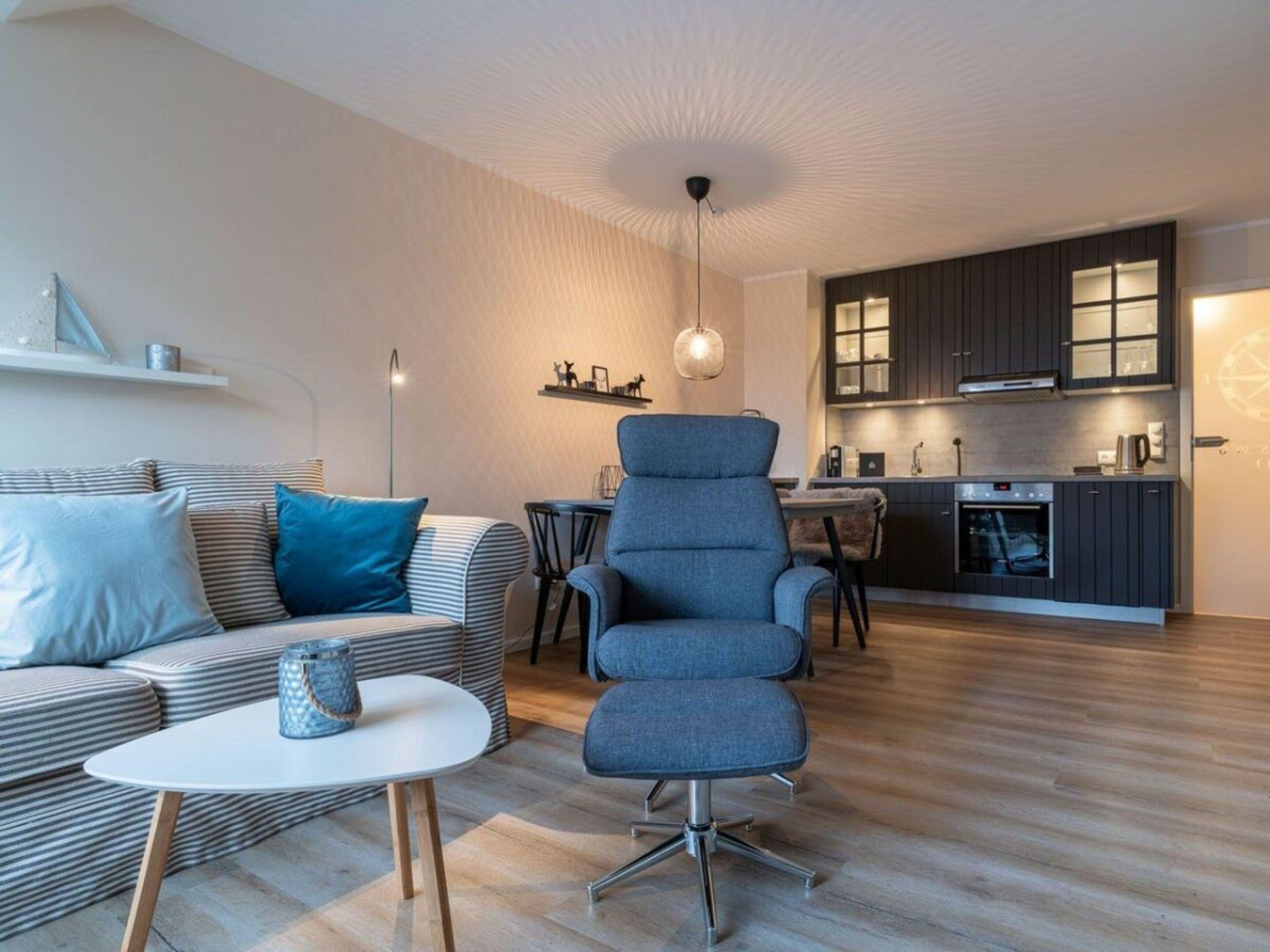 Een moderne woonkamer met een bank, een salontafel en een blauwe fauteuil. Er is een keuken met donkere kastjes en een eettafel.