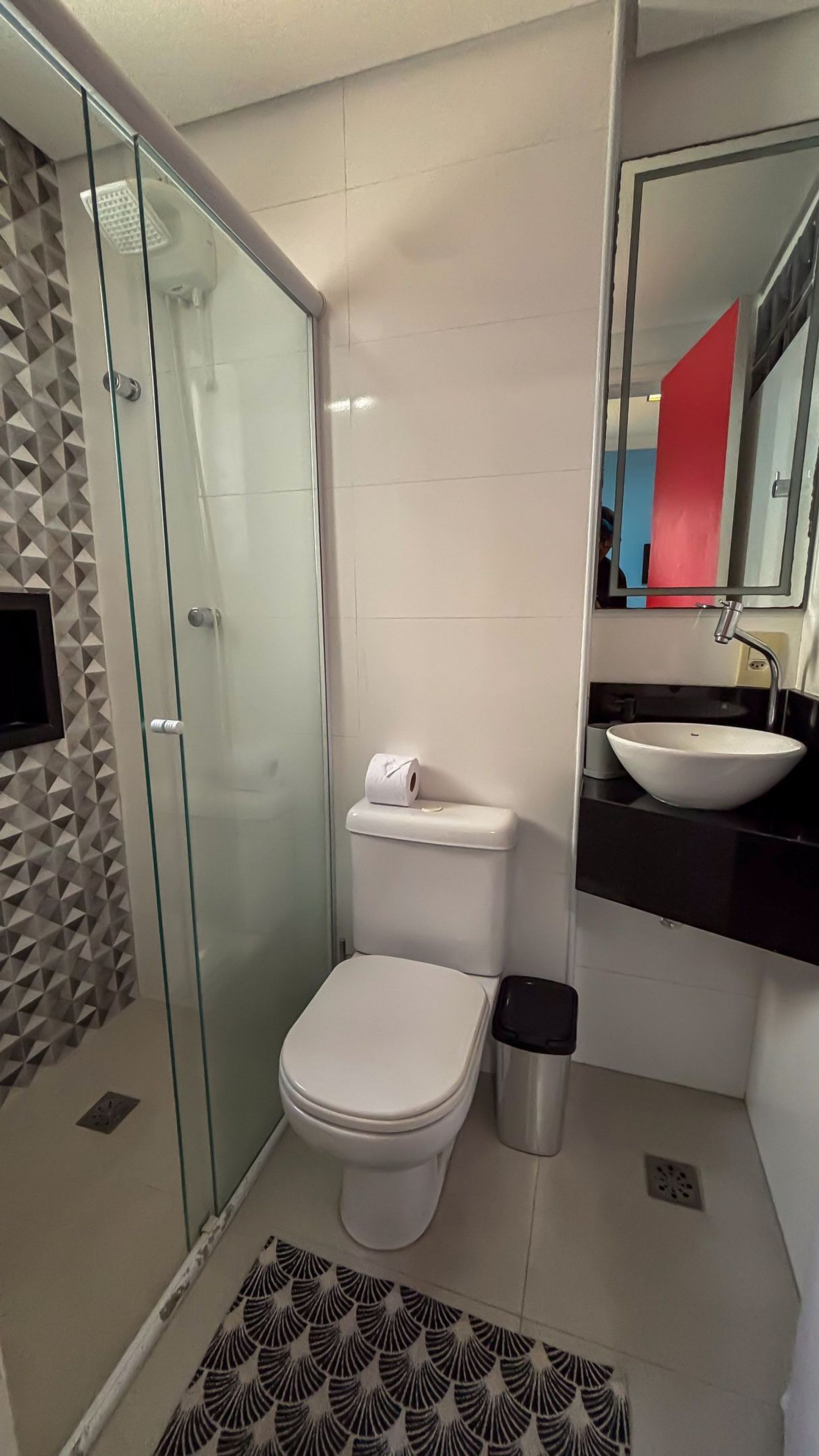 Ein modernes Badezimmer mit einer gläsernen Duschkabine, einer weißen Toilette, einem schwarzen Mülleimer und einem weißen Waschbecken mit Spiegel darüber.