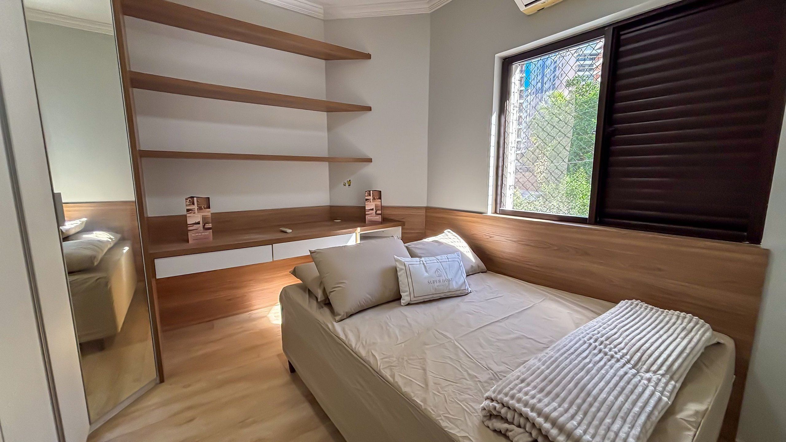 A bedroom with a bed, a built-in shelf unit, a window with blinds, and a view of greenery outside.