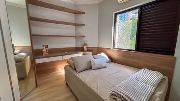 A bedroom with a bed, a built-in shelf unit, a window with blinds, and a view of greenery outside.