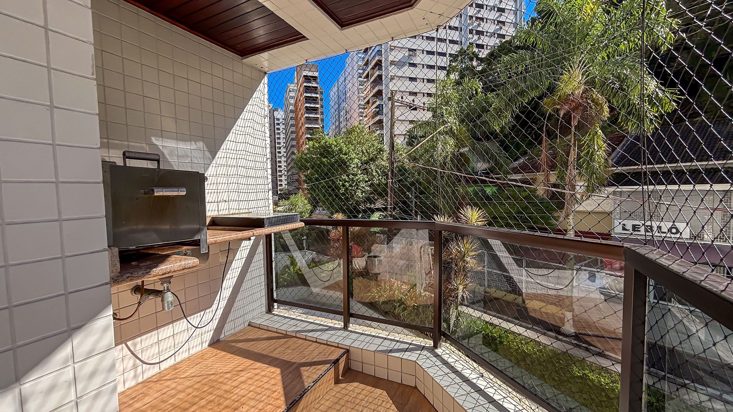 A balcony with a grill, tiled walls, and a view of a cityscape with high-rise buildings and greenery.
