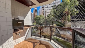 A balcony with a grill, tiled walls, and a view of a cityscape with high-rise buildings and greenery. - SHB - 3 Suítes a 2 min praia (Guarujá)