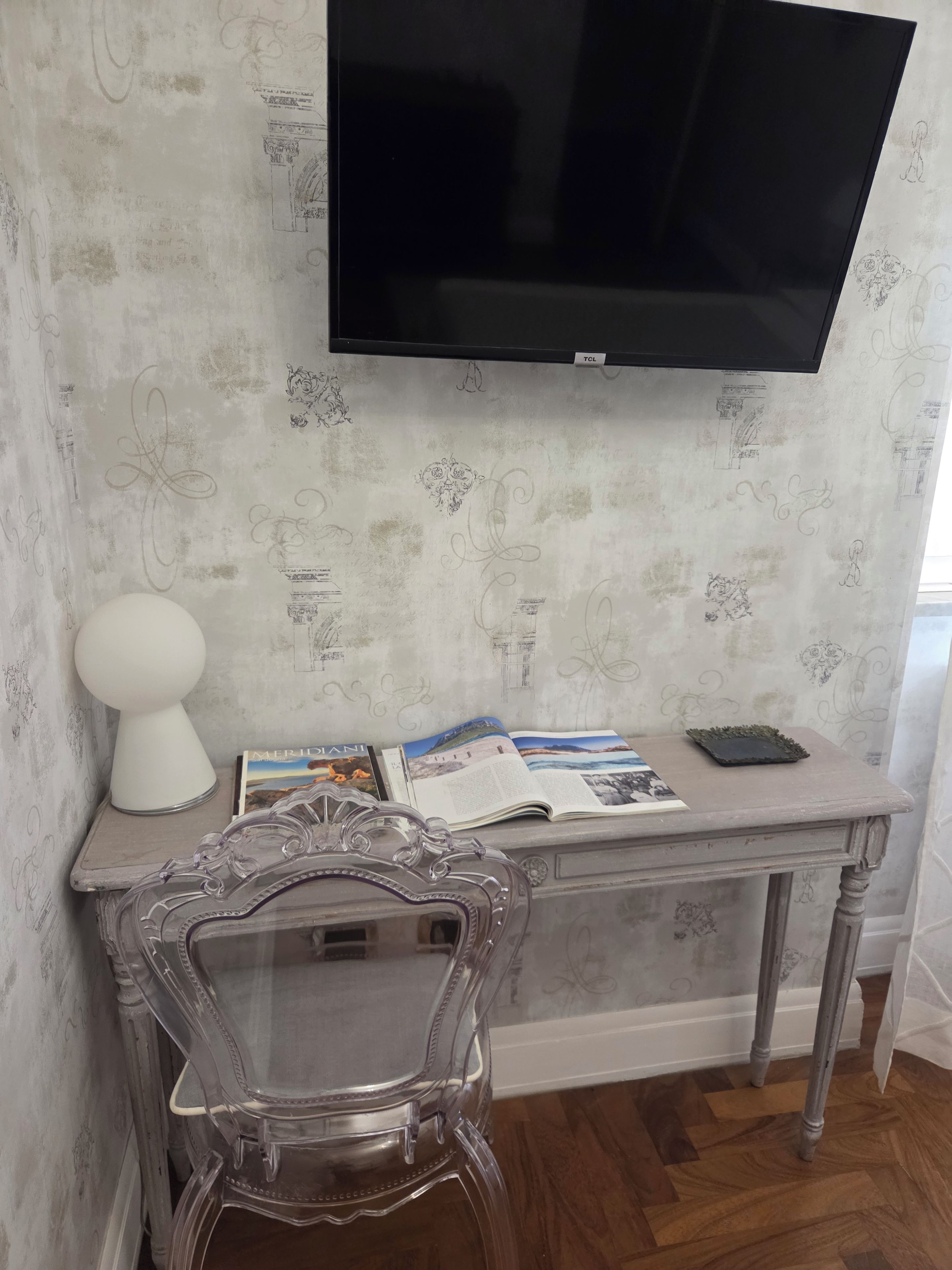 Quarto com televisão de tela plana na parede, uma cadeira de vidro transparente, luminária branca e mesa com revistas e um suporte.