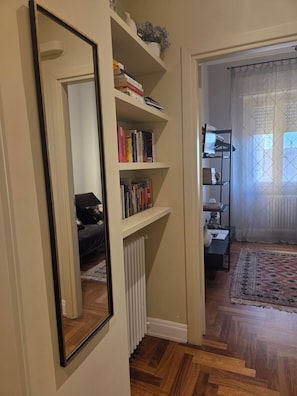 Un pasillo con un radiador blanco, suelo de madera y una estantería llena de libros.