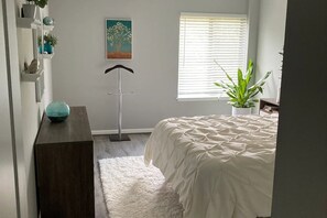 Ein Schlafzimmer mit einem Bett, einem weißen Teppich, einer Pflanze, einem Bild an der Wand und einem Fenster mit Jalousien.