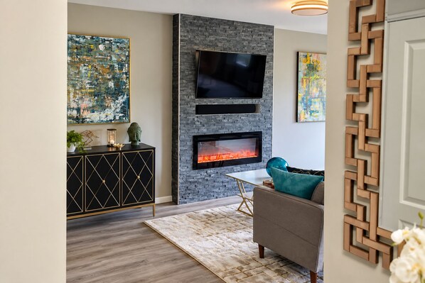 A living room with a fireplace, a flat-screen TV mounted on a stone wall, a sofa, and a decorative cabinet. - Luxury Stay-Free Parking-Nearby DC-Wifi-Pool-Gym (Falls Church)