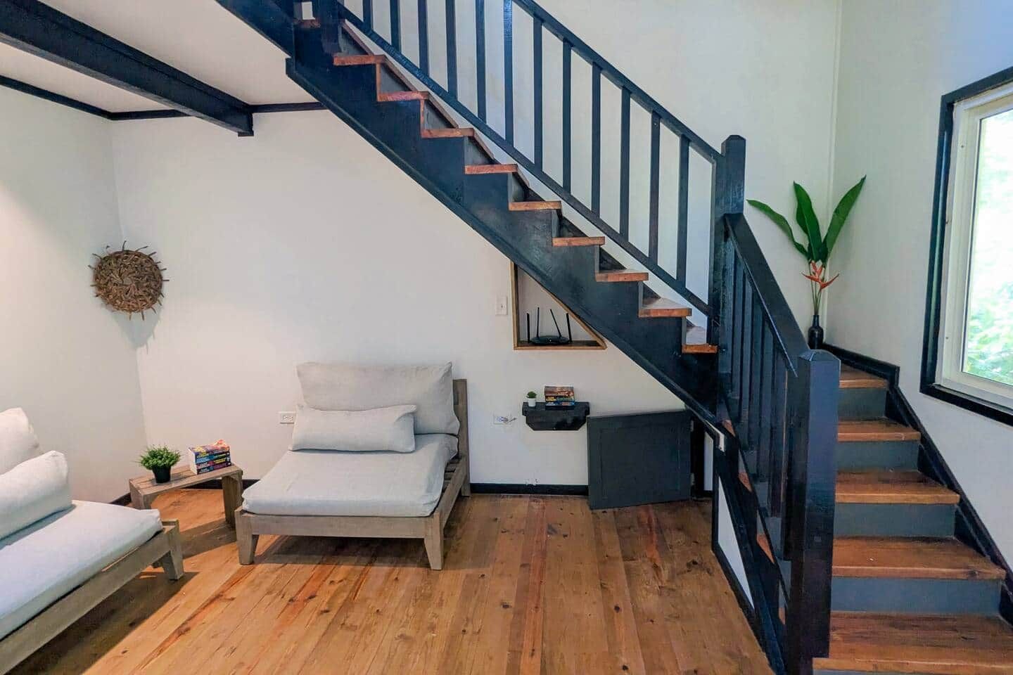 A modern room with a wooden floor, a bed, a small table with a plant, a wall-mounted shelf, and a staircase with black railings.