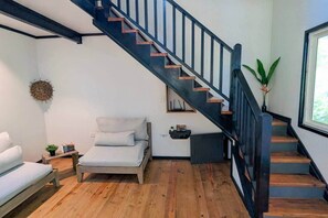 A modern room with a wooden floor, a bed, a small table with a plant, a wall-mounted shelf, and a staircase with black railings.