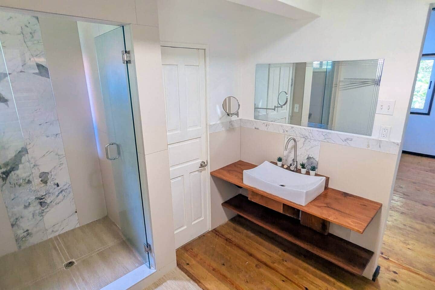 A bathroom with a glass shower enclosure, a white sink with a wooden countertop, and a mirror above the sink.